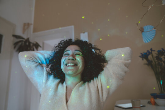 Happy woman with hands behind head and eyes closed sitting under astro lights at home - Powered by Adobe