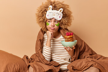 Upset thoughtful woman with curly hair stays in bed under blanket holds bowl of tasty appetizing pancakes with surup applies green hydrogel patches under eyes looks away keeps hand under chin