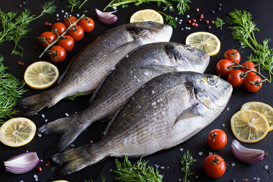 raw dorado fish or sea bream on black background with with lemon, vegetables and herbs