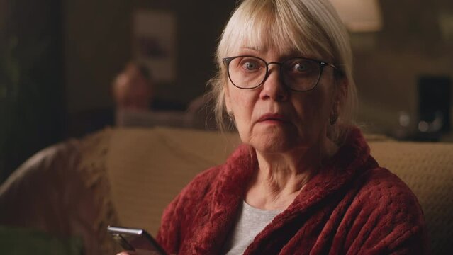 Confused aged woman sitting on the couch in living room while surprising to receive a shocking and terrible sms on a smartphone