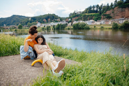 Woman looking and embracing daughter sitting at riverbank