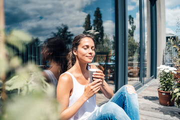 Smiling woman with eyes closed holding disposable coffee cup sitting in front of glass window at balcony