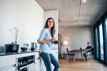 Smiling woman with bowl standing in kitchen with freelancer using laptop at home