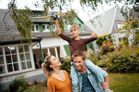 Happy woman looking at son holding airplane sitting on man shoulders at backyard