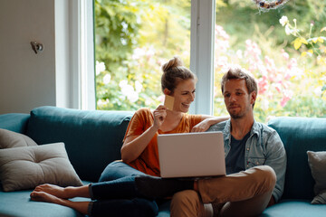 Cheerful woman holding credit card sitting by boyfriend using laptop on sofa in living room