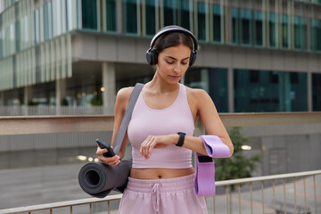 Outdoor shot of sporty fit woman poses with sport equipment checks time on smartwatch uses...
