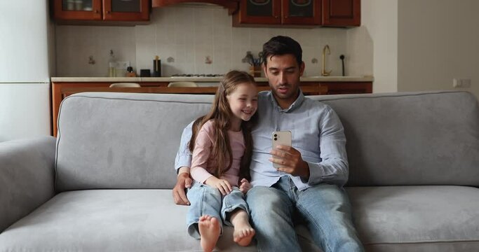 Young father his little 6s adorable daughter relax on sofa at home with smartphone listen music watch video clip enjoy internet content. Family leisure using modern tech, technology overuse concept