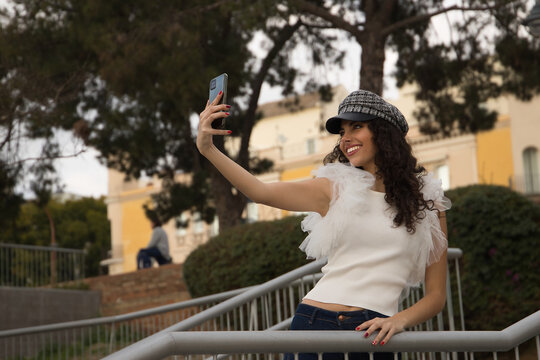 Young and beautiful woman, brunette, with curly hair, wearing white shirt, jeans and cap, taking pictures of herself with her cell phone. Concept beauty, fashion, model, trend, selfie, photos.