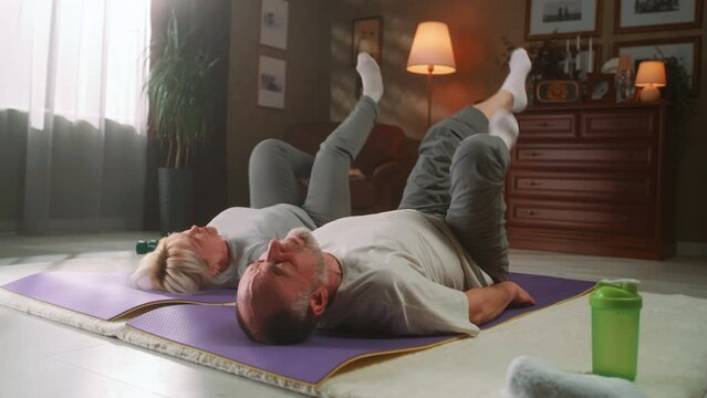 Senior spouses in sportswear actively working out together on a fitness mat at home and exercising on leg muscles