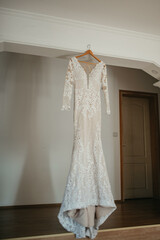 Wedding dress hangs on a chandelier in a white room.