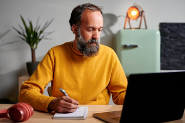 Middle-aged, bearded, focused man in yellow sweatshirt, taking some notes in a notebook while using...