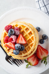 Homemade waffles with berries, powdered sugar and maple syrup on white plate, gray background.