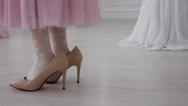 Close-up of children's feet in big women's beige shoes high heels. Little girl walks in mom shoes