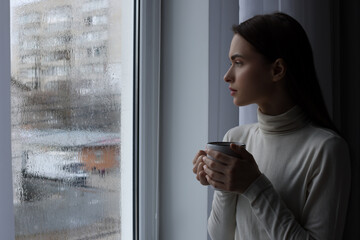 Melancholic young woman with drink looking out of window on rainy day, space for text. Loneliness concept