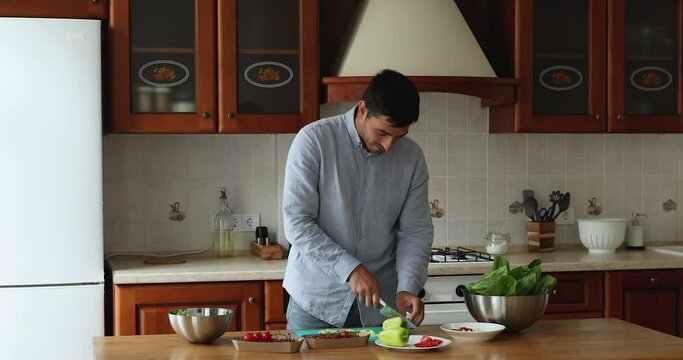 Young handsome man enjoy cooking alone in kitchen, cutting paprika for vegetarian salad, prepare romantic dinner, move to music looks carefree. Healthy habit lifestyle, homemade food recipes concept
