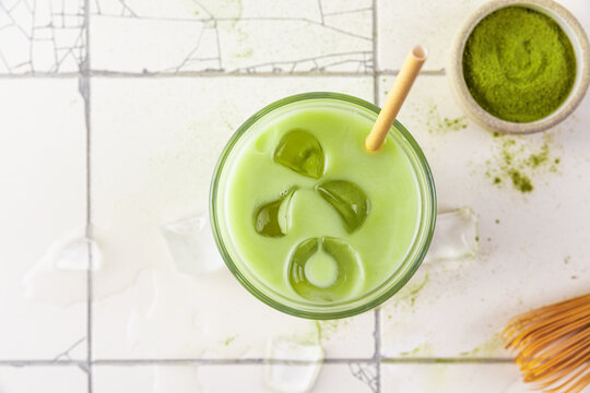 Glass of iced green matcha latte with a straw on white tile background. top view