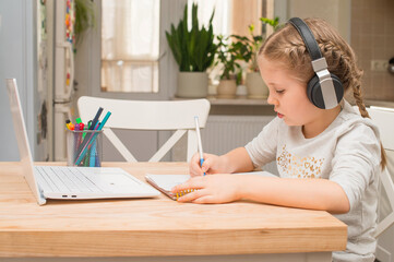 Little schoolgirl wearing wireless headphones makes videocall using web cam and notebook writing notes learning school subject distantly. Remote self-education concept.