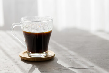a transparent cup of hot brew black coffee with morning shadows and sun rays through the window on gray table background, close up. Morning coffee concept