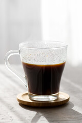 a transparent cup of hot brew black coffee with morning shadows and sun rays through the window on gray table background