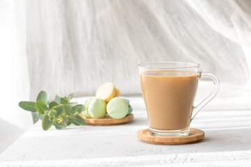 a transparent cup of coffee with milk with macaroons and eucalyptus leaves at morning shadows and sun rays through the window on gray table background. Morning coffee concept