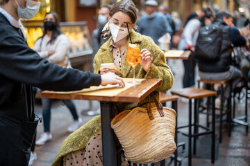 Waiter brings a menu for a young female customer at outdoor cafe. Woman wearing medical mask visiting public place