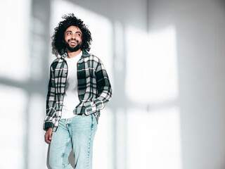 Handsome smiling hipster  model.Sexy unshaven Arabian man in summer stylish shirt and jeans clothes.Fashion male with long curly hairstyle posing near wall in studio.At sunny day, shadow from window