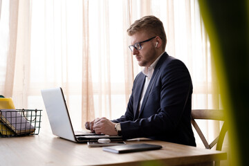Blond caucasian young businessman wearing suit and glasses with headphones working with white laptop at desk in office or at home near big window. Man using computer