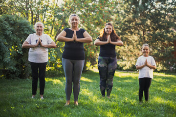Few generations of females from one family standing together in Sukhasana pose on backyard full of...