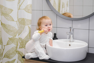 A toddler sitting on the washbasin cabinet and brushing teeth, showing independence, learning new skills. Daily morning hygiene routine. Personal and teeth hygiene.