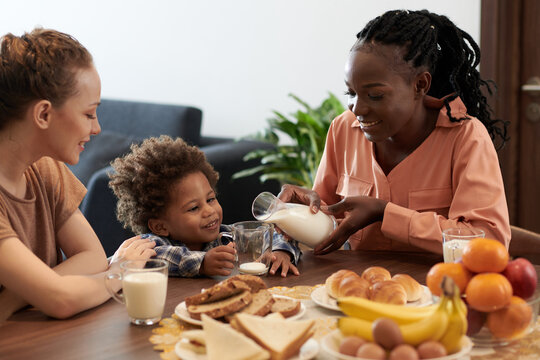 Smiling Young Mother Pouring Milk In Cup For Her Baby Boy At Family Breakfast