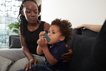 Worried mother looking at son using inhaler when having asthma attack at home