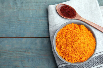 Bowl of saffron powder and spoon with dried flower stigmas on light blue wooden table, flat lay. Space for text