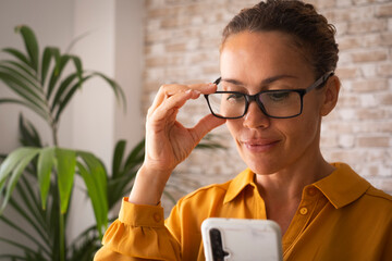 Confident portrait of young pretty adult woman smiling and using mobile phone connection at home or office. Attractive female people wearing eyeglasses and reading cellular device social media