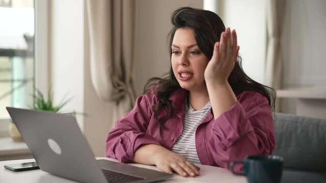 Young overweight woman looking at laptop and talking while sitting at table in home office spbd. 4k Close view of beautiful European female looks at screen and talks with smile, gestures and sits at