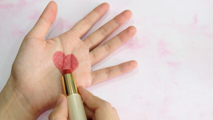 Hand holding a tube of lipstick, drawing a heart shape in the center of a palm.