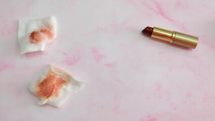 Two pieces of cotton pads with red stain from wiping off lipstick. With a tube of lipstick on the...