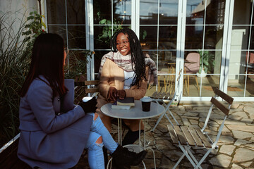 Two multiracial friends sitting on the terrace of cafe on cold weather and having girly talks.