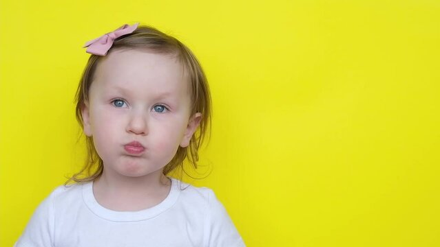 Funny little girl kid puffed out her cheeks and then blows them off while looking at camera on yellow background.