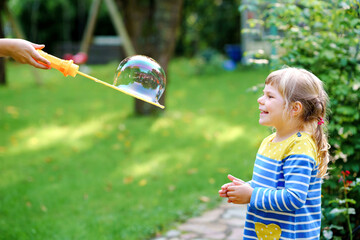 Happy little blonde preschool girl having fun with blowing soap bubble blower. Cute child playing...