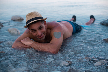 Man in hat lies on beach Dead sea. Gray mud smeared on hand in shape of heart. Salt and mud healthy...