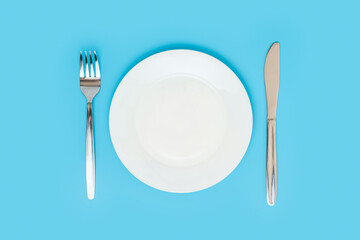 Empty white ceramic plate with a fork and a table knife on a blue background, top view
