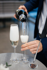 The groom's hand pours champagne from a bottle into two glasses to celebrate after the wedding ceremony closeup