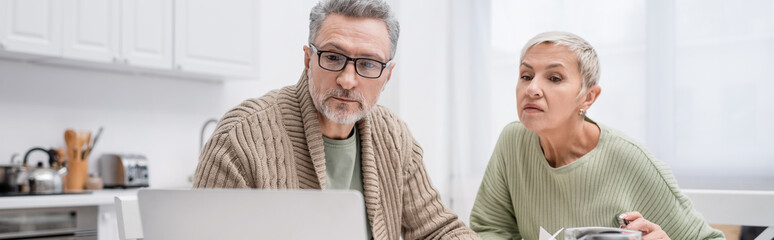 Mature man looking at blurred laptop near wife in kitchen, banner.