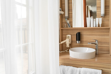 Stylish bright clean bathroom in hotel room with window or door, face care set, curtain, towels, sink, modern faucet, wooden furniture, hair dryer and mirror on bright day. Travel, vacation, holidays