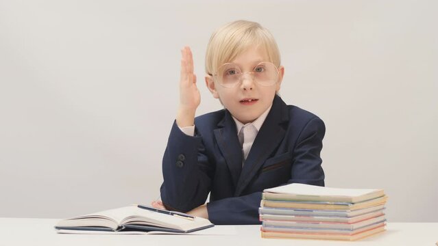 Schoolboy sits at desk with his hand raised wants to answer teachers question. Boy in school uniform at desk next to stack of notebooks. Diligent student.