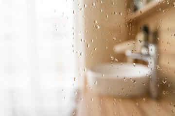 Water drops on wet glass shower door in hotel bathroom with blured bokeh window, sink, faucet and wooden furniture on sunny morning or day. Travel, holiday, vacation, interior design, body care