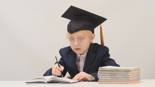 Little boy in school uniform and students hat sits at desk and writes in notebook. Child as university professor.