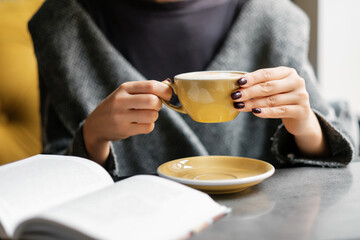 The girl drinks coffee from a yellow mug and reads a book in a quiet cafe at a table by the window.