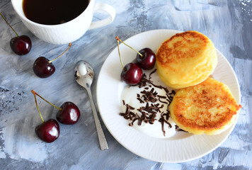 syrniki with sour cream, chocolate chips and cherries on plate and cup of coffee and teaspoon