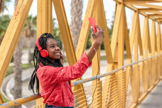portrait of a smiling young african american woman with braids listening to music with her red headphones and taking a selfie with her cell phone.